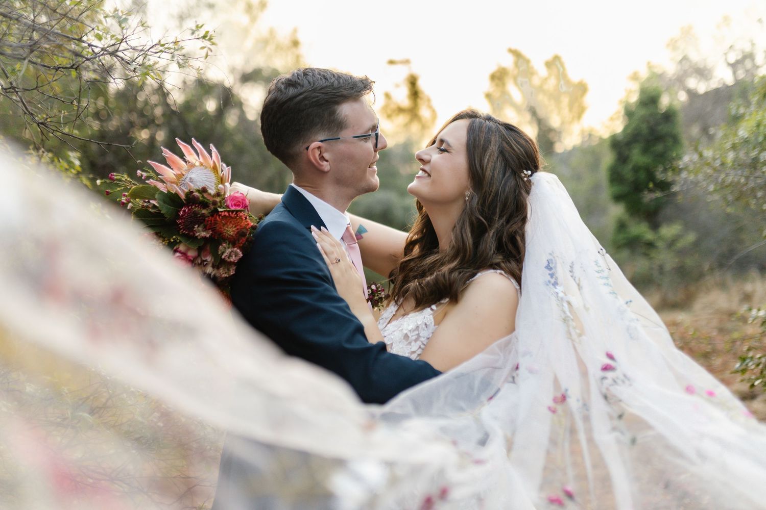 A romantic wedding couple embraces in a scenic outdoor setting with a flowing white veil catching the sunlight.