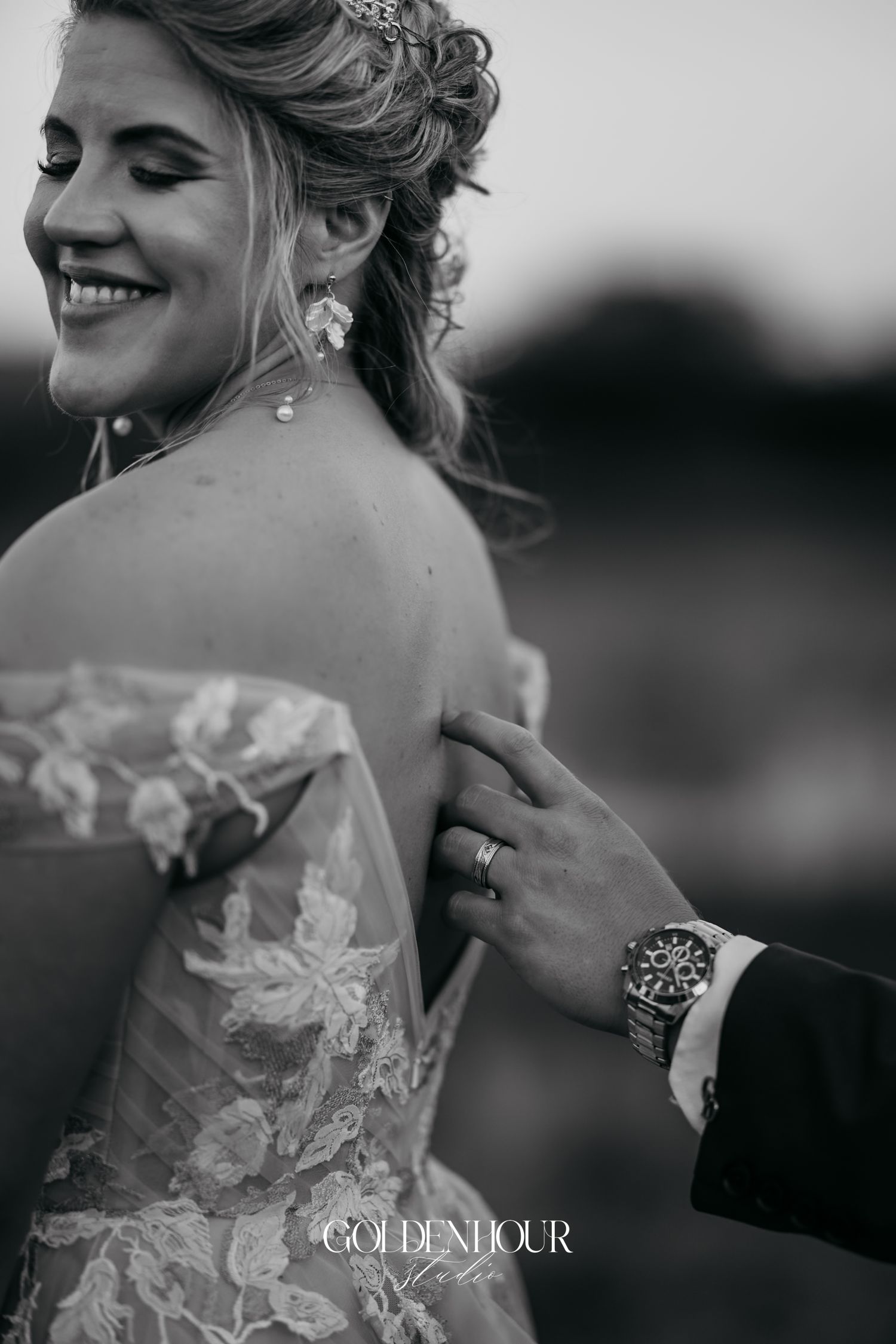 Black and white photo of smiling bride in lace gown as groom adjusts her dress outdoors.