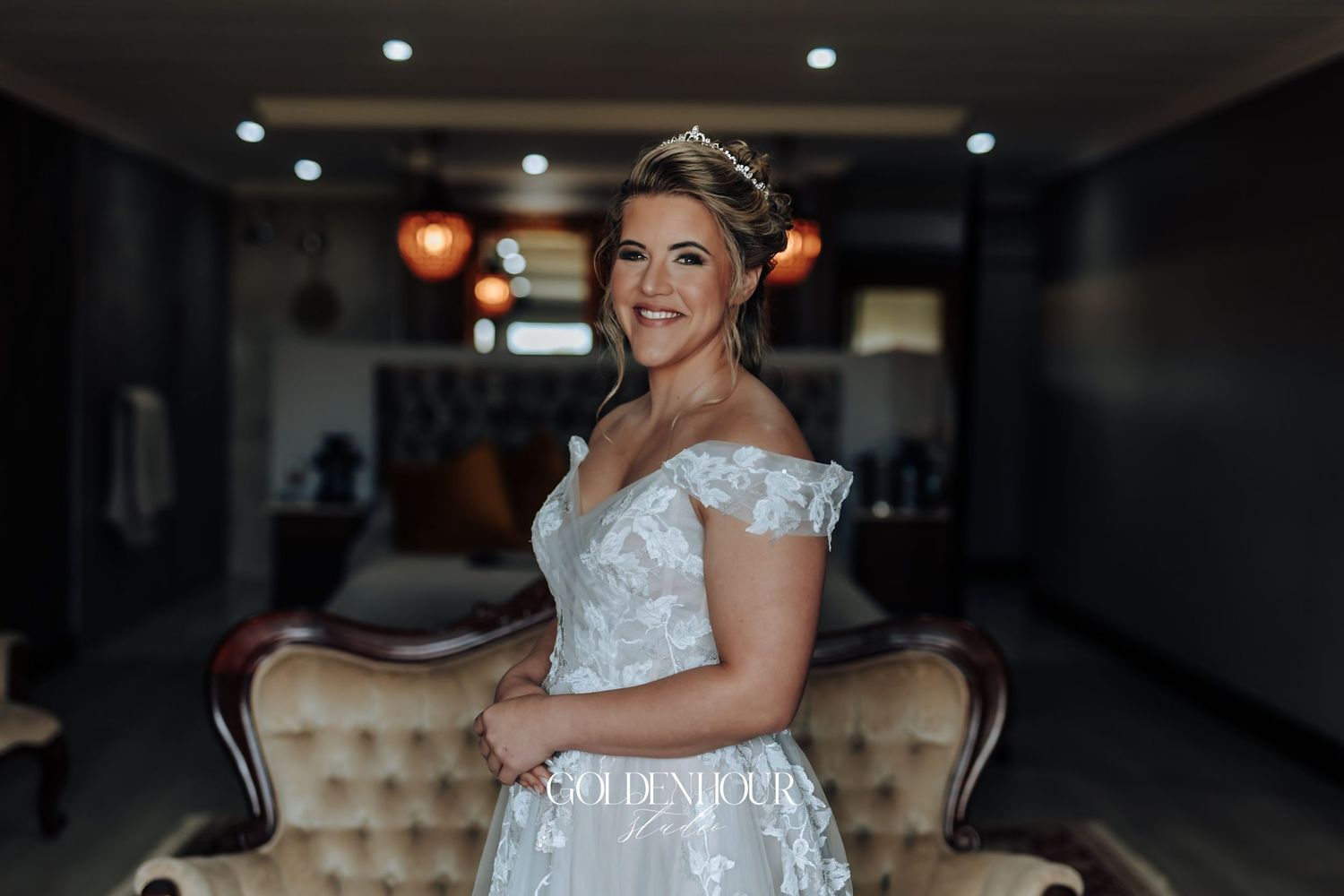 Radiant bride in off-shoulder lace wedding gown wearing tiara in elegant interior.