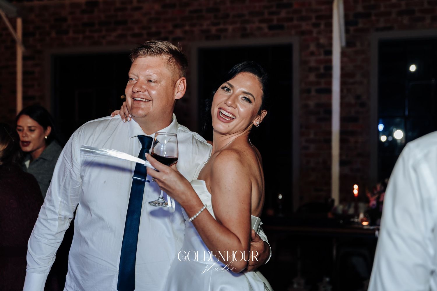Joyful bride in white dress laughs with guest holding wine glass at wedding reception.