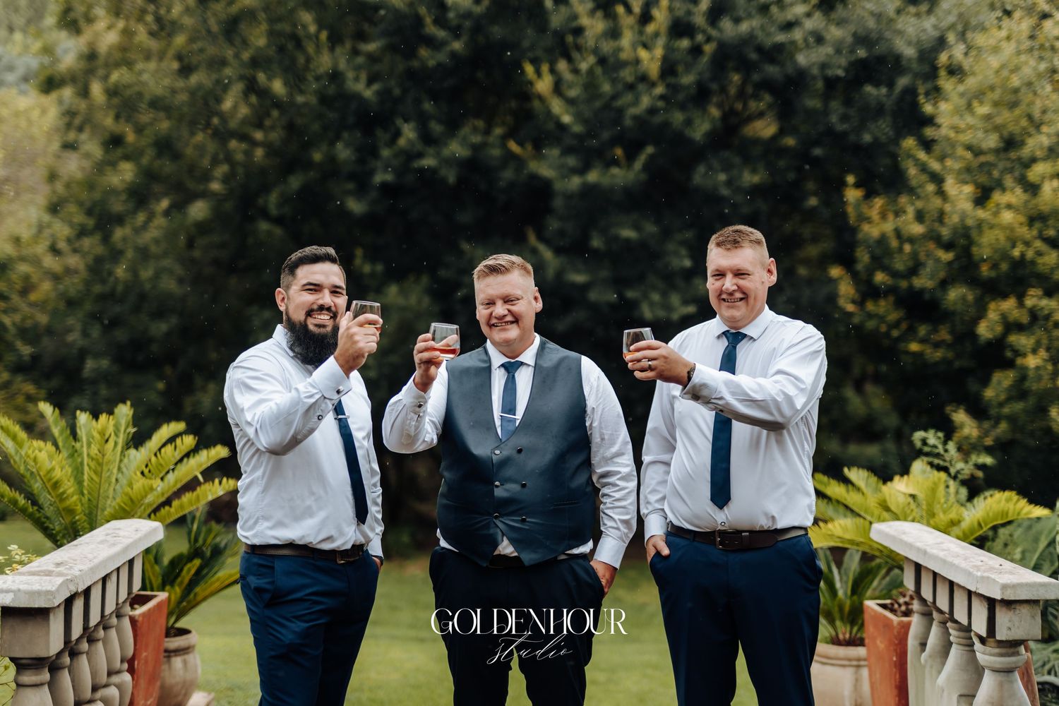 Three groomsmen in white shirts and navy ties toasting drinks outdoors at a wedding venue surrounded by lush greenery.