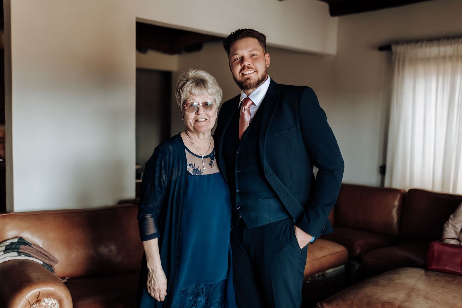 Two people in formal attire standing together in front of a brown leather couch in a room with natural lighting.