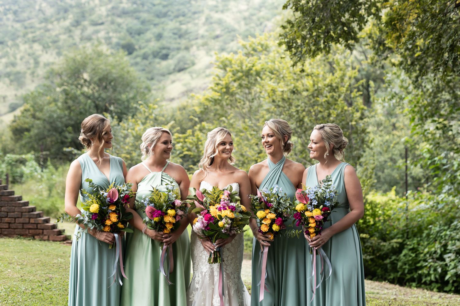 Bridal party wearing sage green dresses holds vibrant sunflower bouquets in an outdoor garden setting.