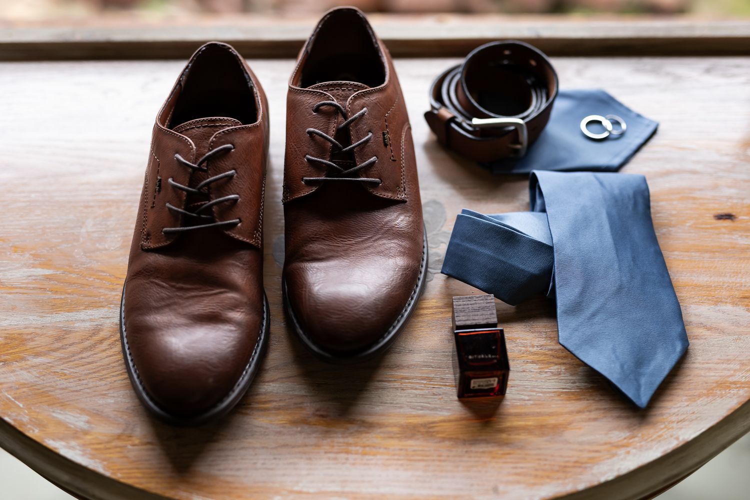 Brown leather dress shoes with blue tie and black belt arranged on wooden surface.