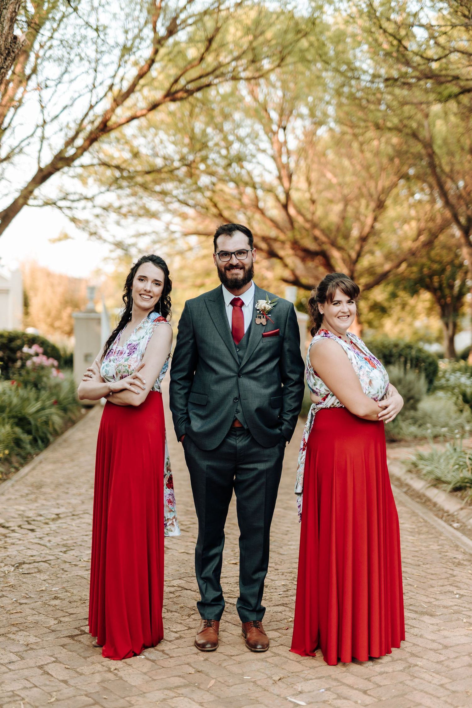 Wedding group stands posed on garden path with bridesmaids in flowing red gowns and groomsman in grey suit.
