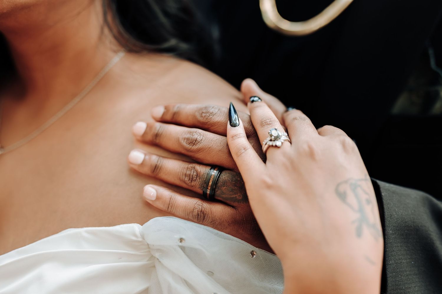 Close-up of couple's hands with wedding rings and tattoos against white dress.