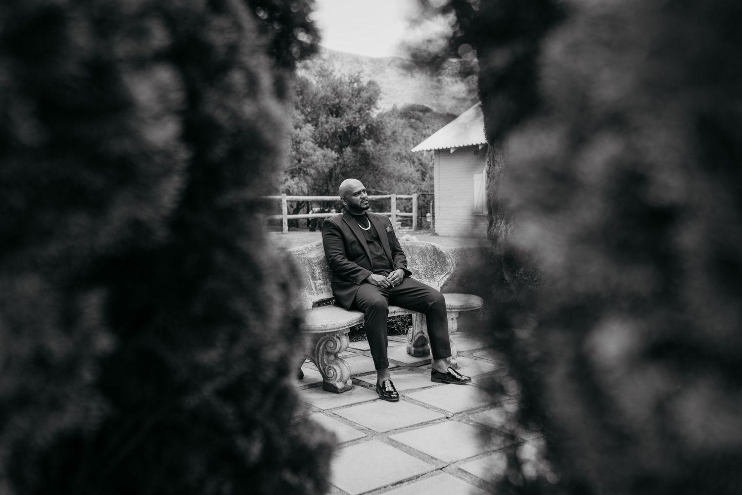 Artistic black and white photo of someone seated on bench in garden through foliage frame.
