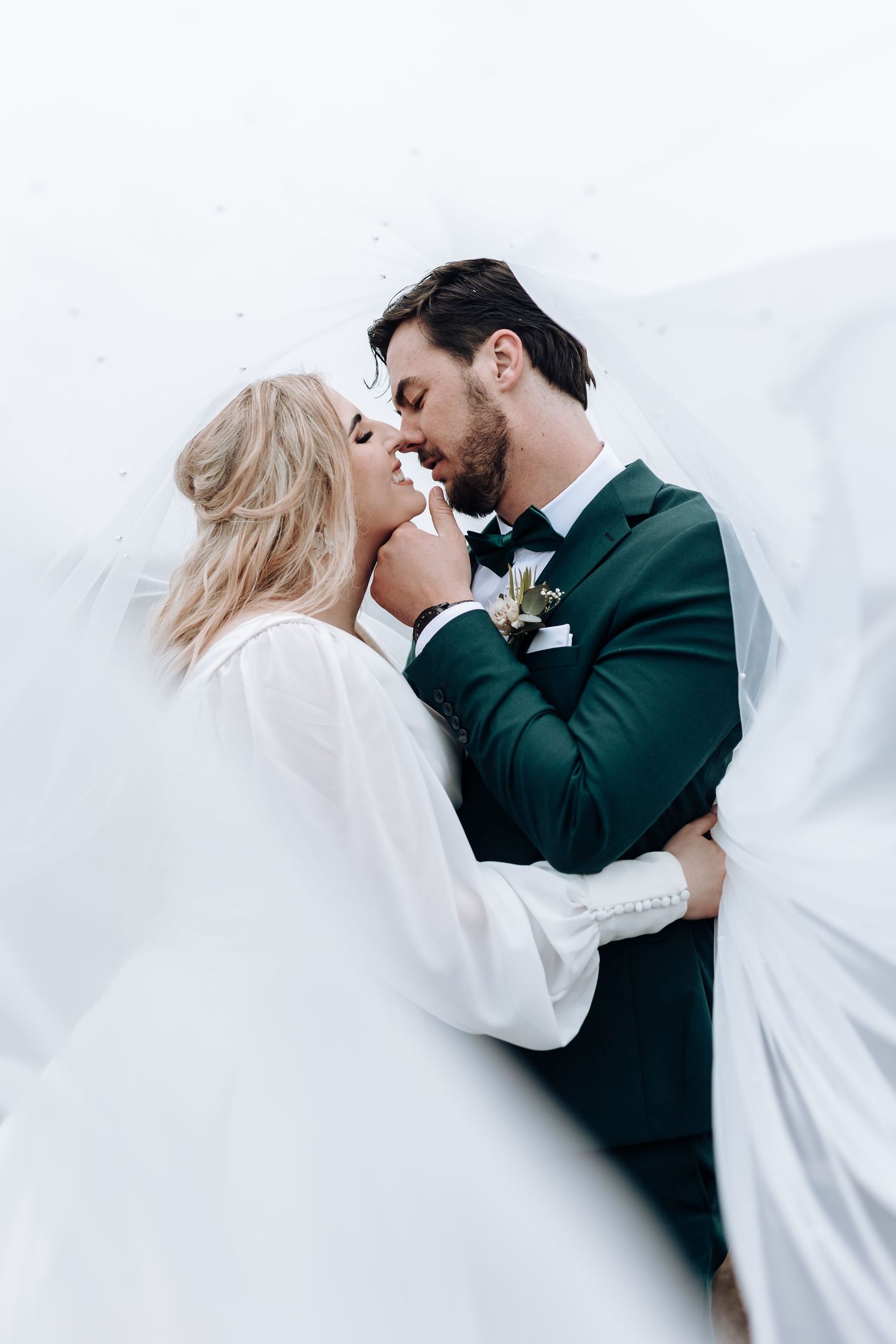 Artistic wedding portrait shows intimate moment between couple with flowing veil and green suit.
