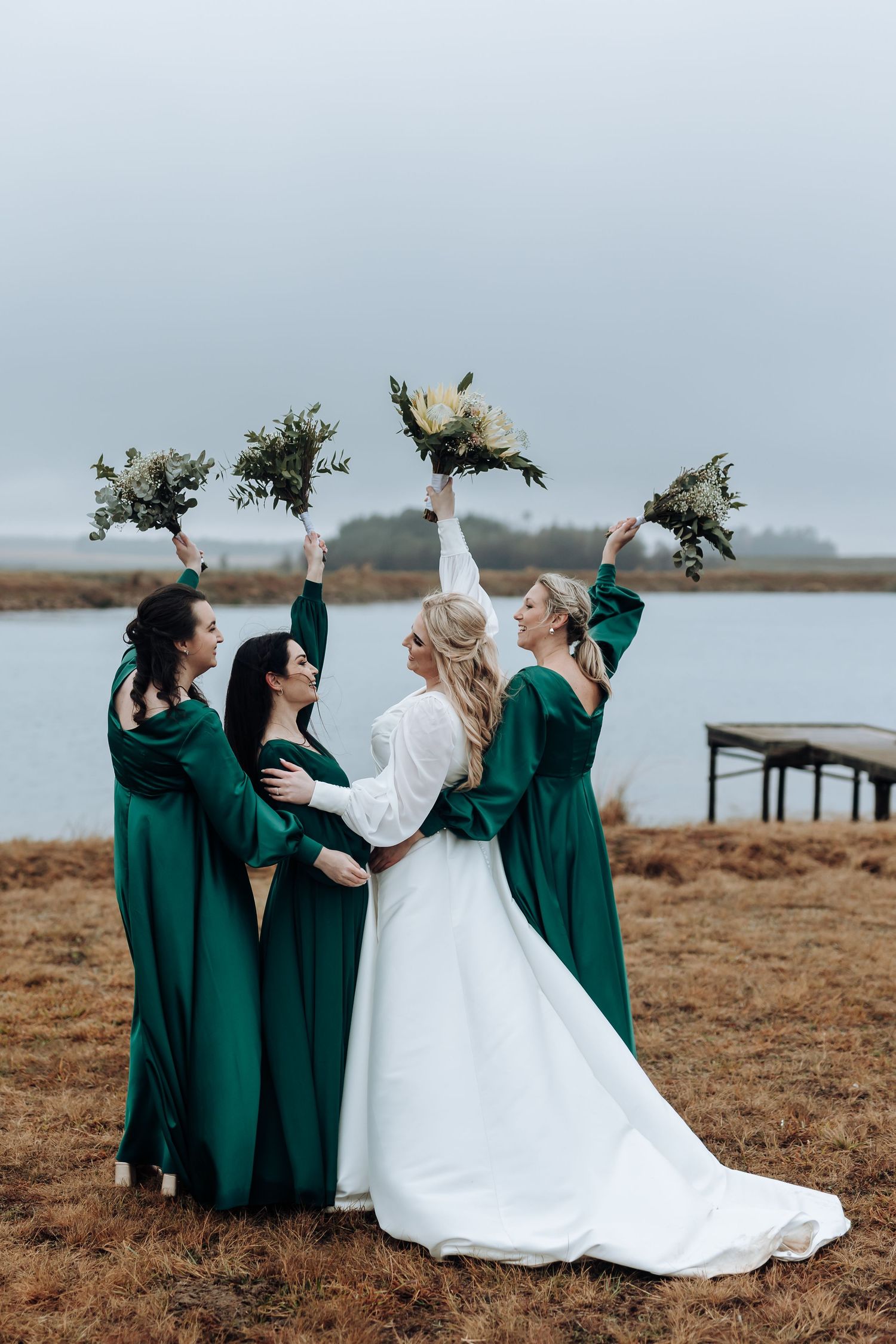 Bridal party in emerald dresses raise bouquets in celebration by the water.