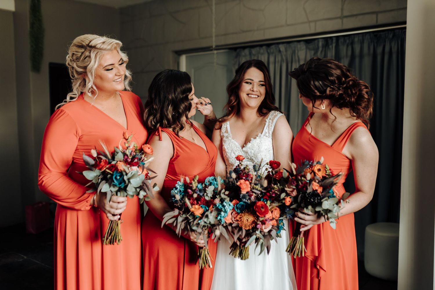 Candid moment of bridal party laughing together while holding bright coral and blue floral bouquets.
