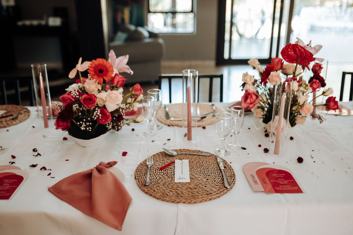 Wedding table setup with coral napkins, rattan chargers, and red rose centerpieces.