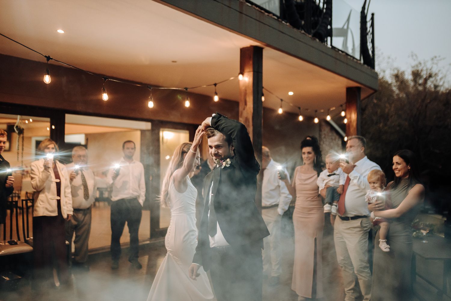 A couple dances surrounded by guests under string lights at an evening wedding reception.