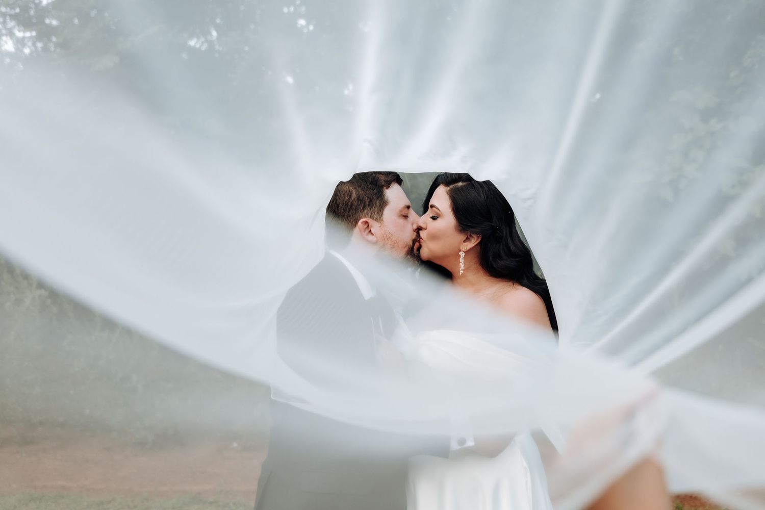 Romantic wedding portrait through flowing white veil with dramatic lighting effect.