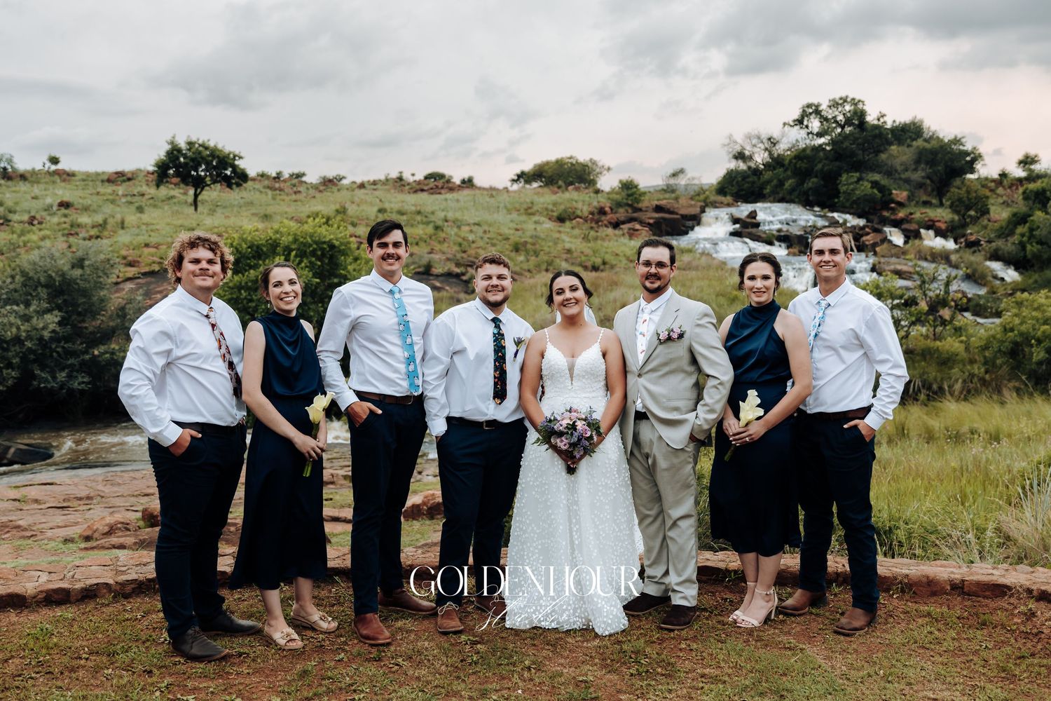 Wedding party lined up for a casual group photo on a grassy hillside.