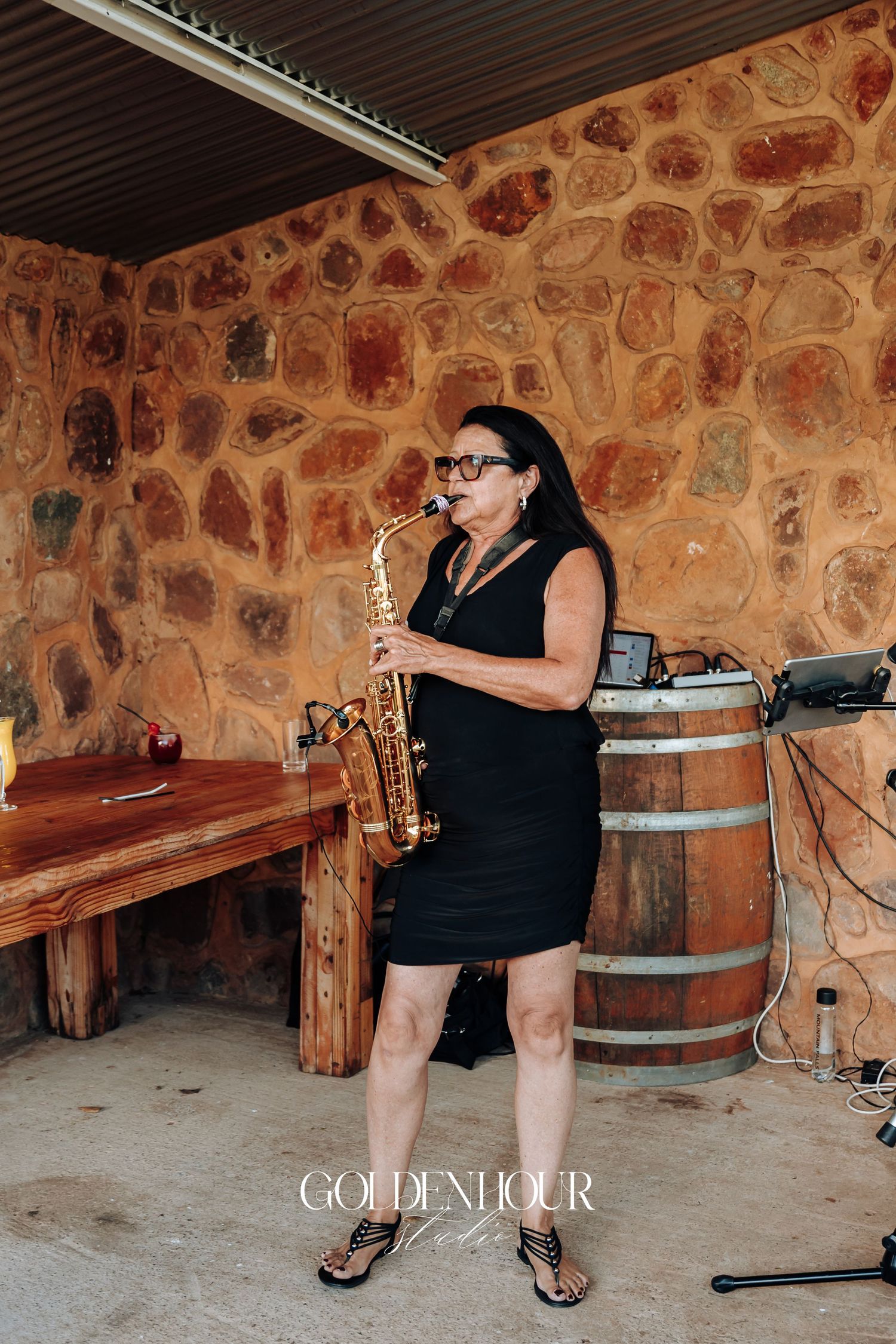 A musician in a black dress plays saxophone against a rustic stone wall backdrop with wooden barrels and benches.