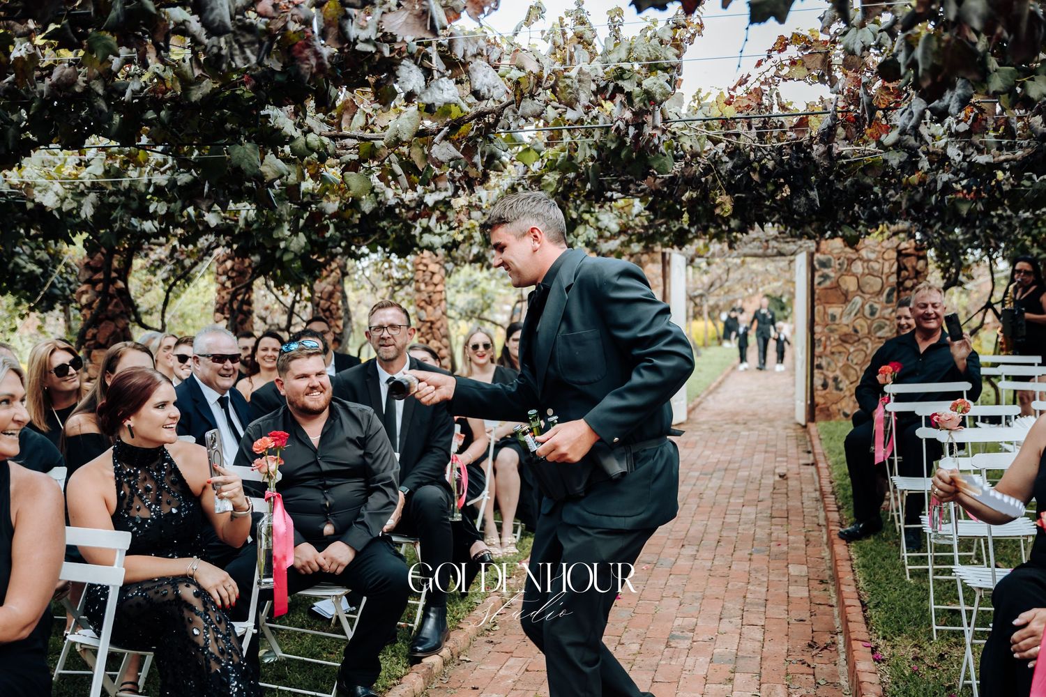 Wedding guest shares a laugh with attendees during outdoor ceremony on brick pathway under floral archway.