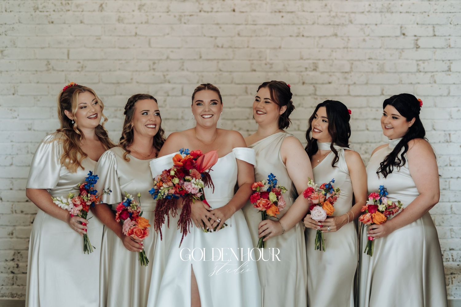A row of people in white dresses stand together holding colorful floral bouquets against a white brick wall.
