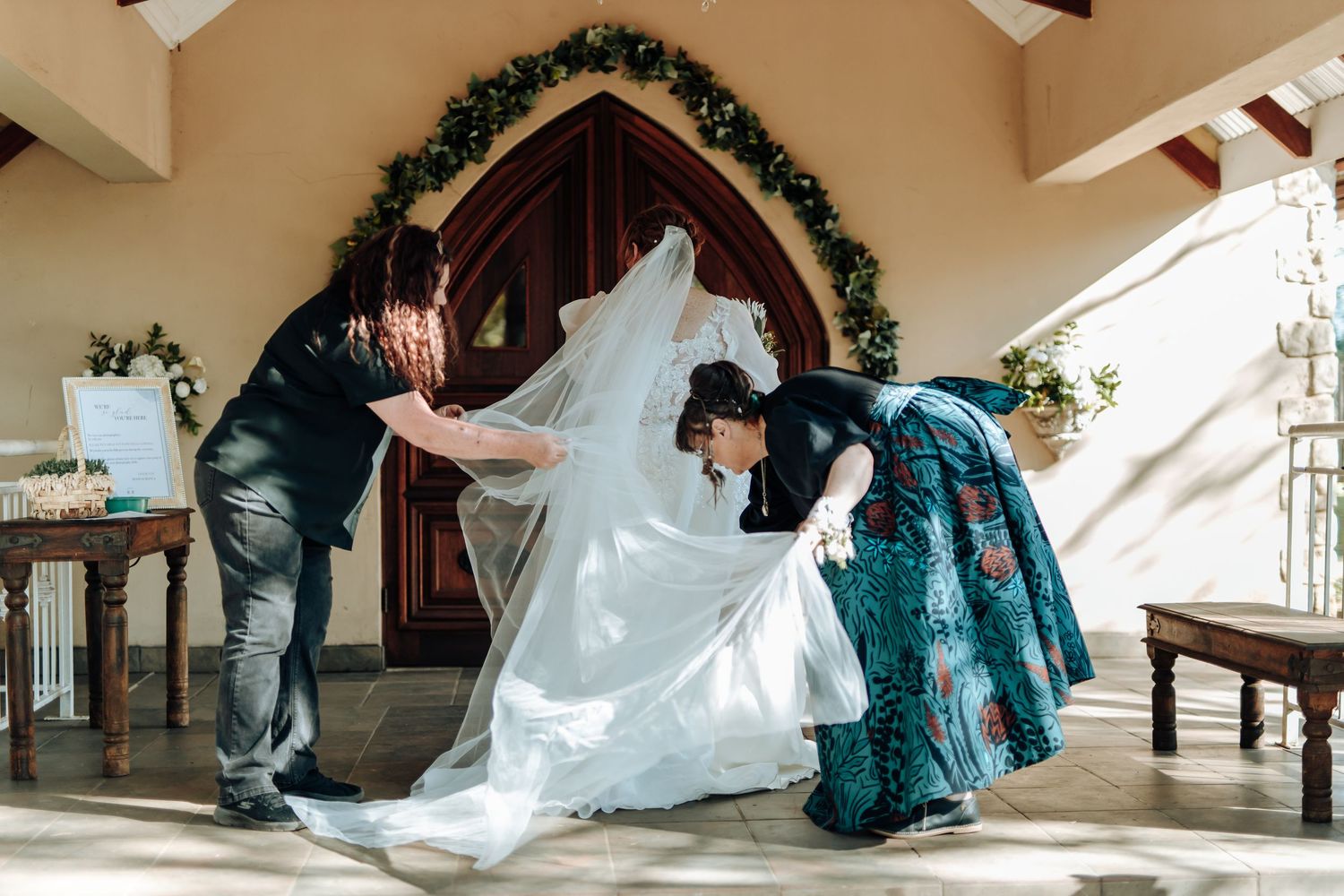 A group adjusts a flowing white wedding dress train on the steps of a chapel entrance decorated with greenery.
