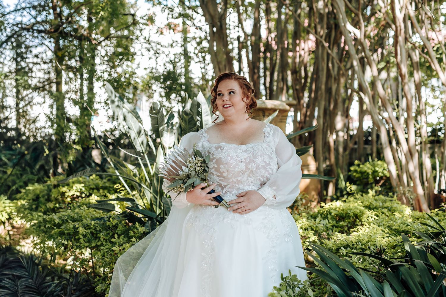 A bride in a flowing white gown stands among Spanish moss draped trees in a romantic garden setting.
