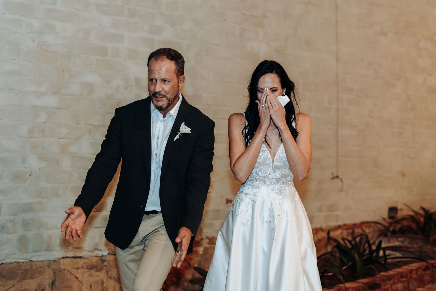 Emotional bride in white lace gown wiping tears beside suited man during wedding ceremony entrance.