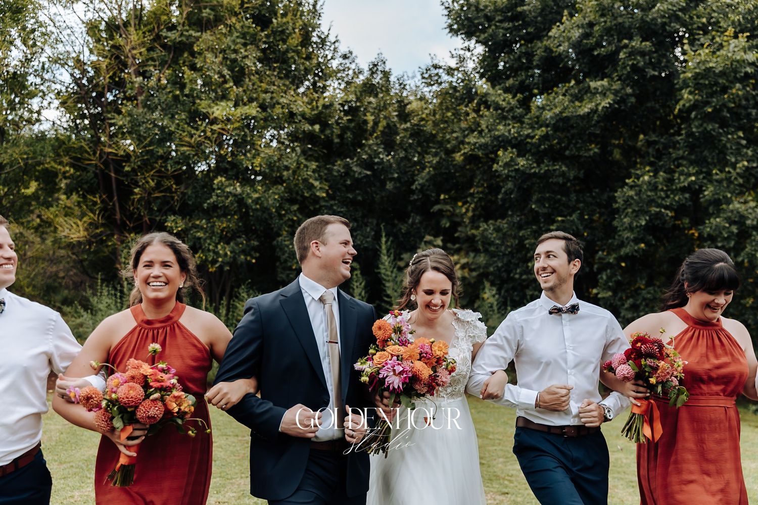 Bride and groom laugh joyfully with wedding party in red dresses on a green lawn.