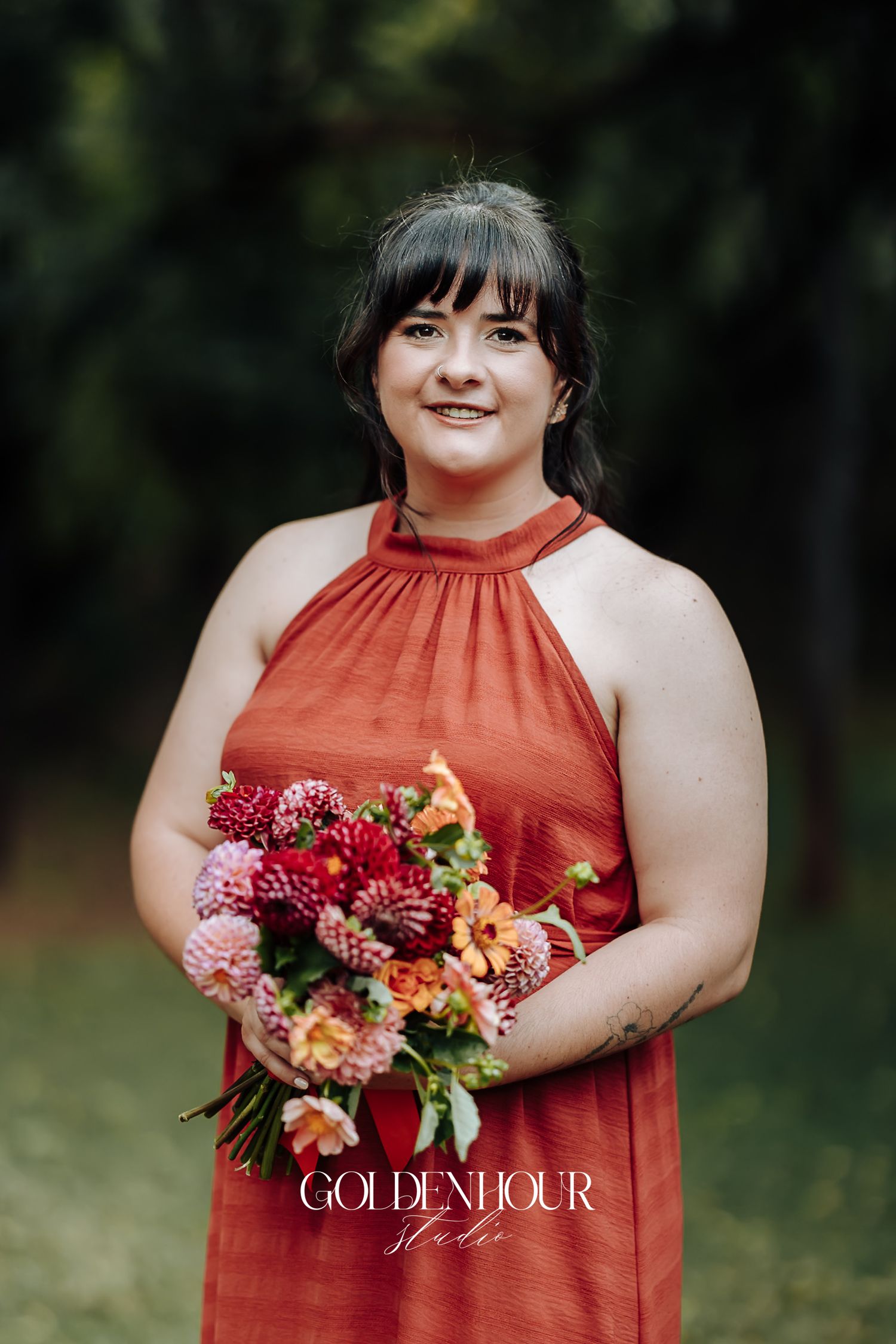 Bridesmaid in rust red halter dress holding vibrant pink and orange floral bouquet outdoors.