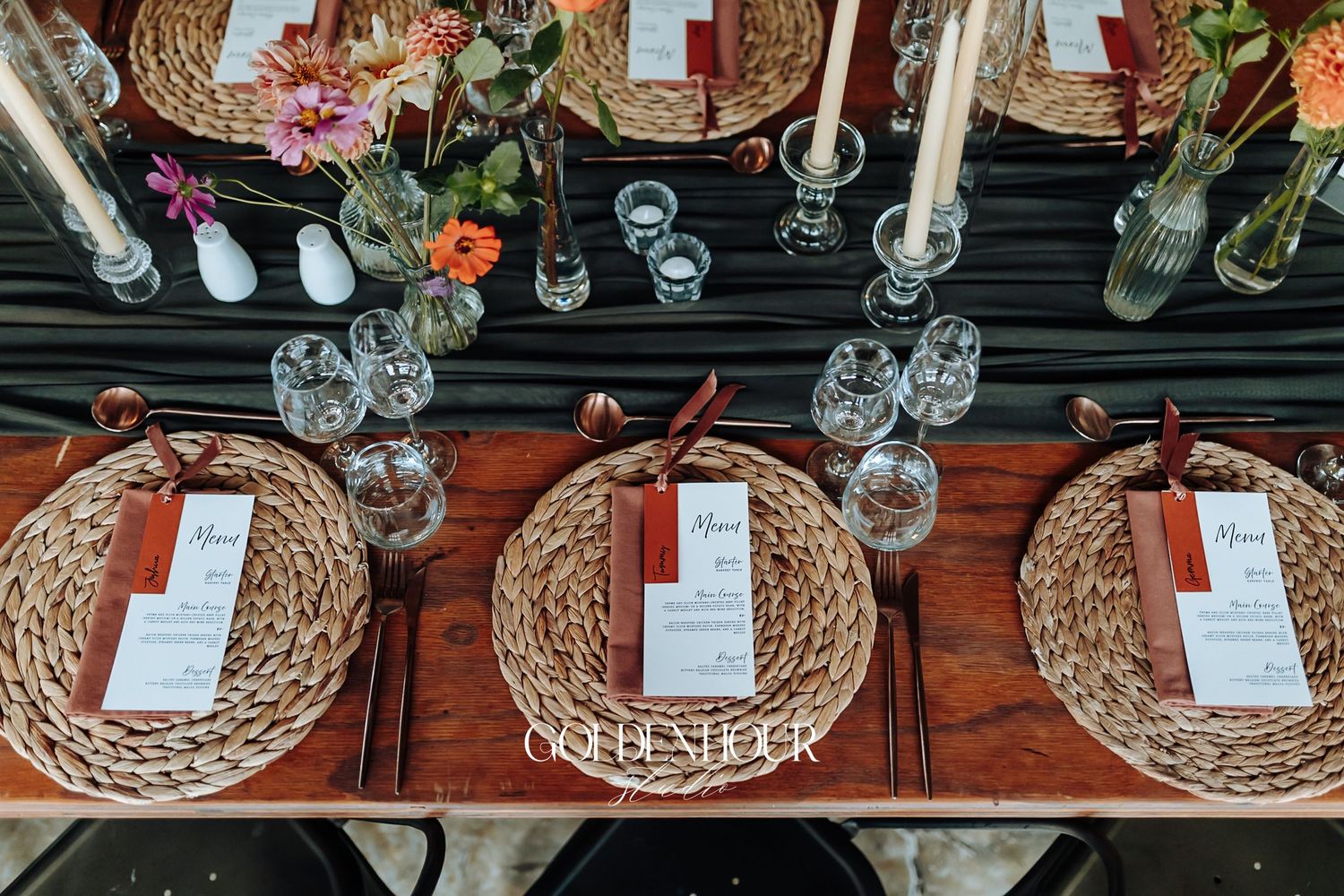 Elegant boho wedding table setting with woven chargers, taper candles, wildflowers, and menu cards on dark linen.