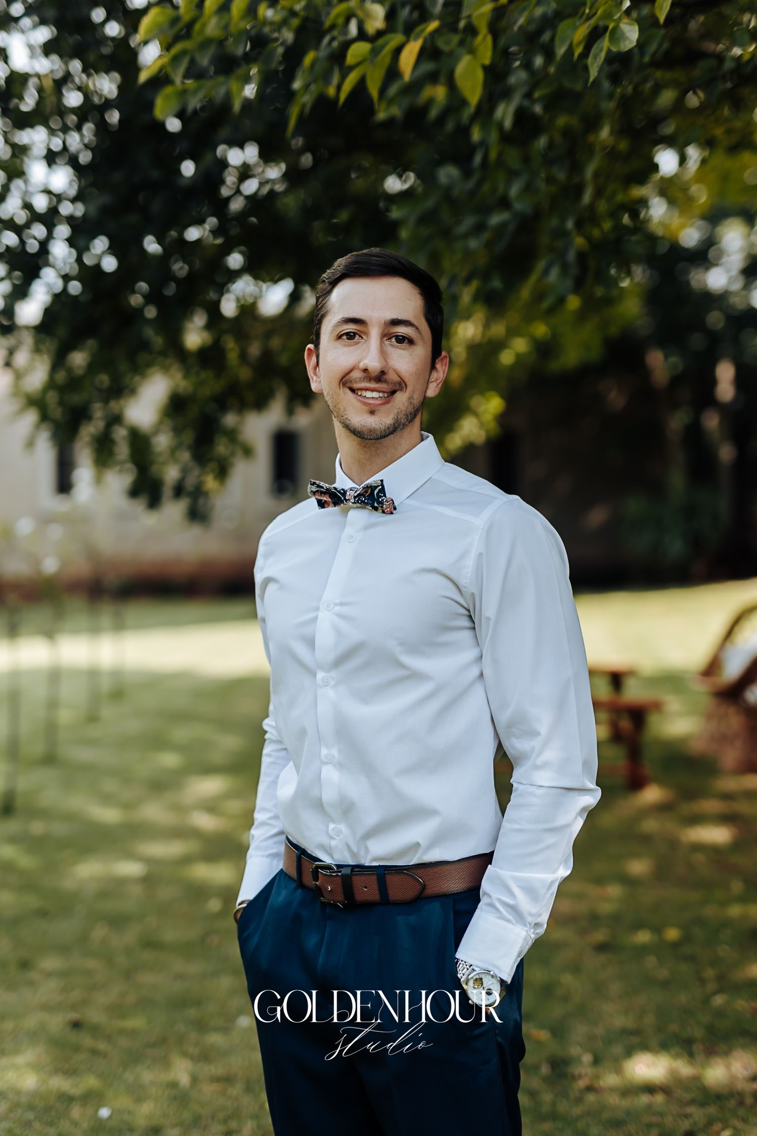 Dapper groomsman in white shirt, navy pants, and floral bow tie smiling outdoors under green trees.