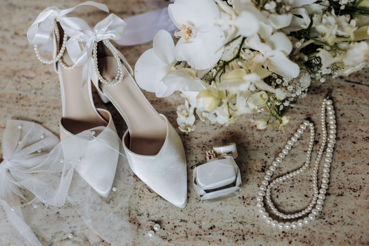 White wedding accessories including pointed heels, pearl necklace, veil and floral arrangement on textured surface.