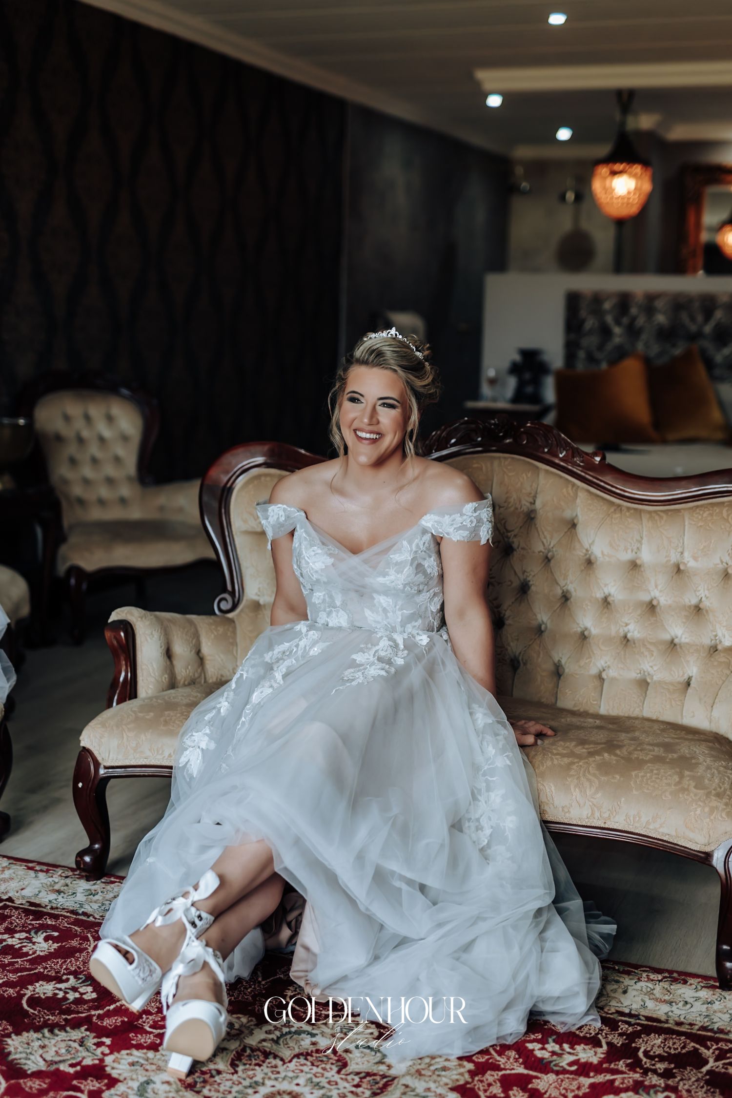 Smiling bride in a white lace off-shoulder wedding dress seated on an elegant vintage sofa with heels.
