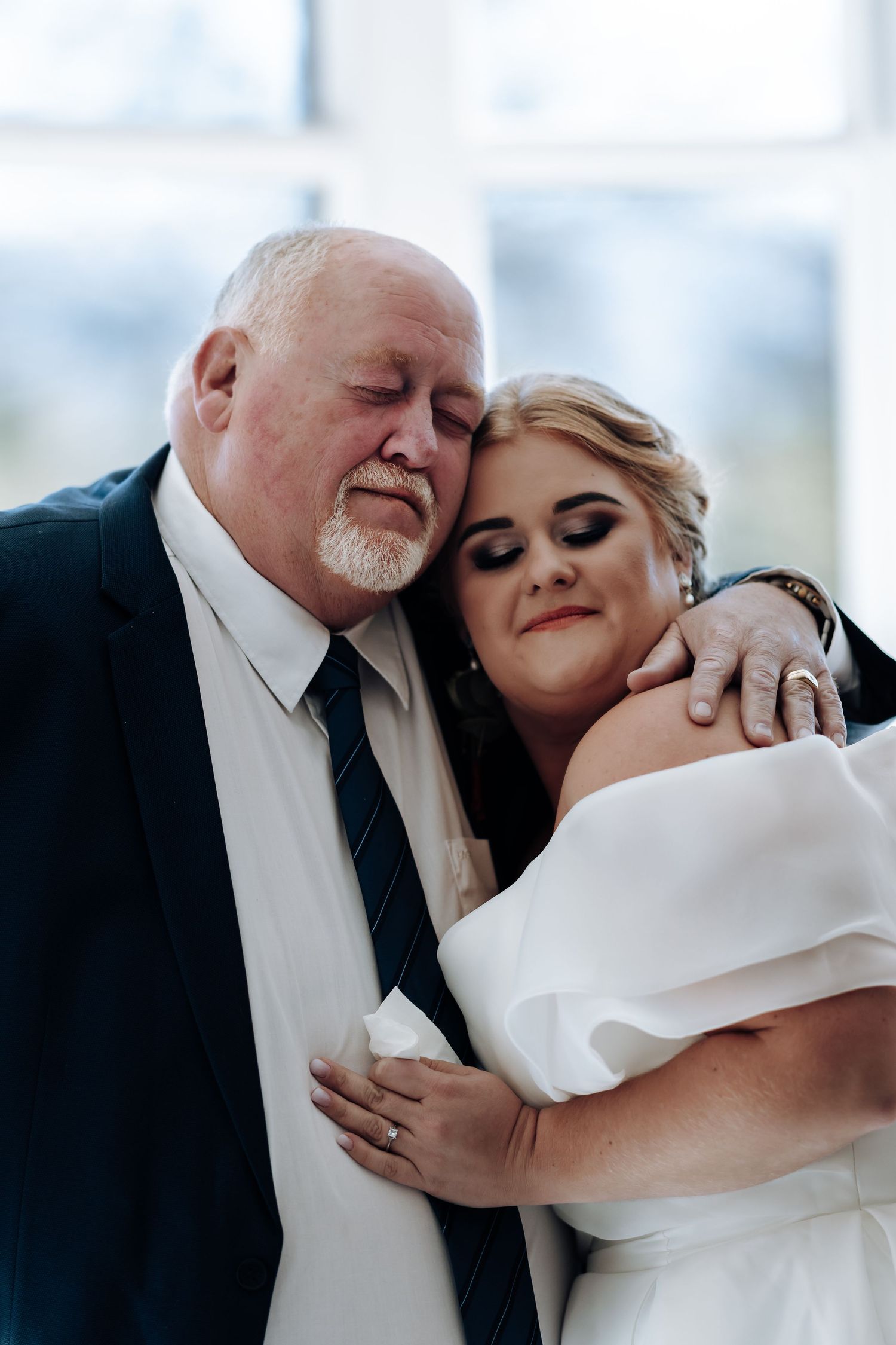 Emotional father-daughter wedding moment, bride in white off-shoulder gown embracing older man in navy suit and tie.