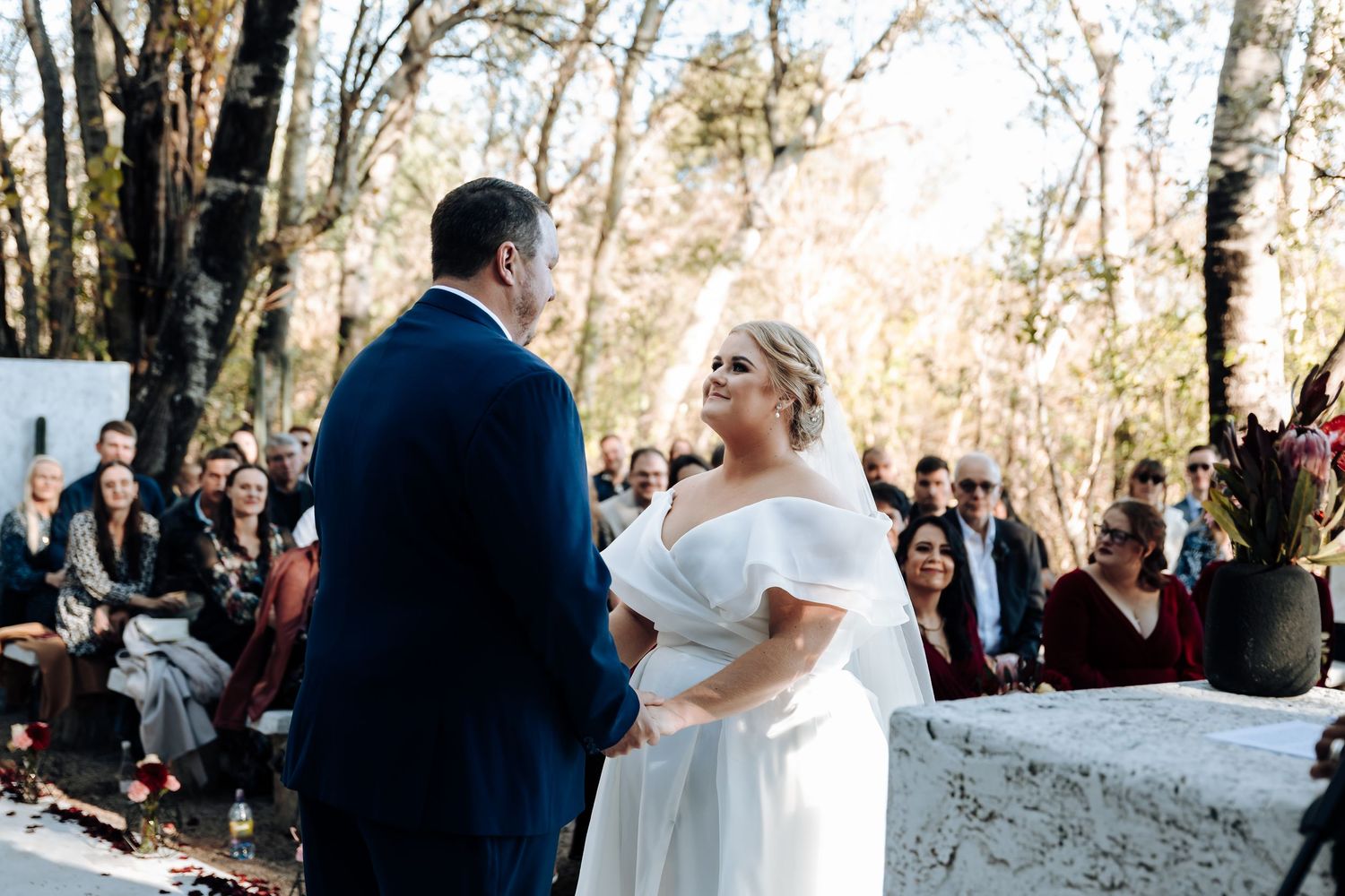 Bride and groom exchange vows at outdoor woodland wedding ceremony with guests seated among trees.