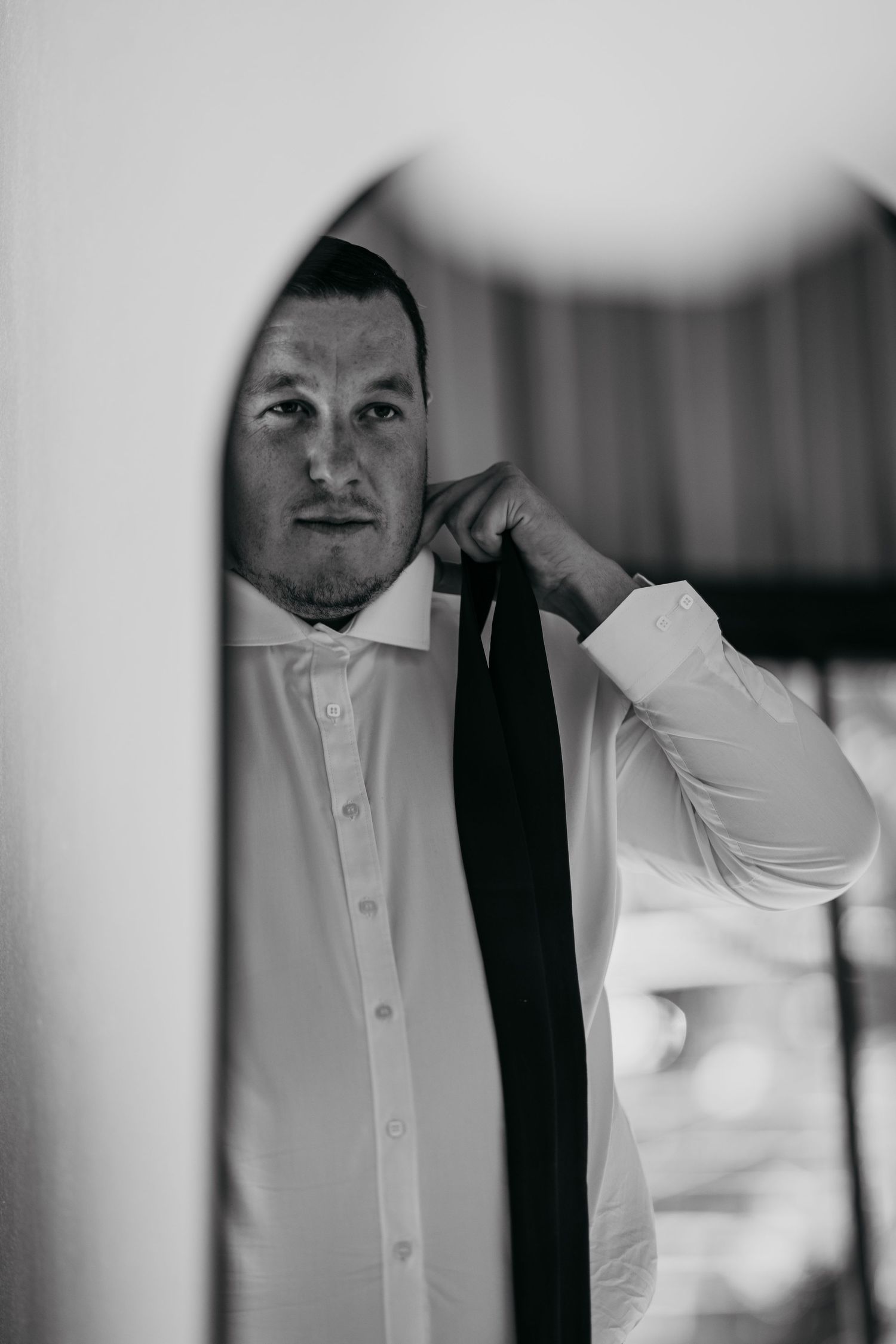 Black and white photo of a groom adjusting his tie in a mirror while getting ready for his wedding.