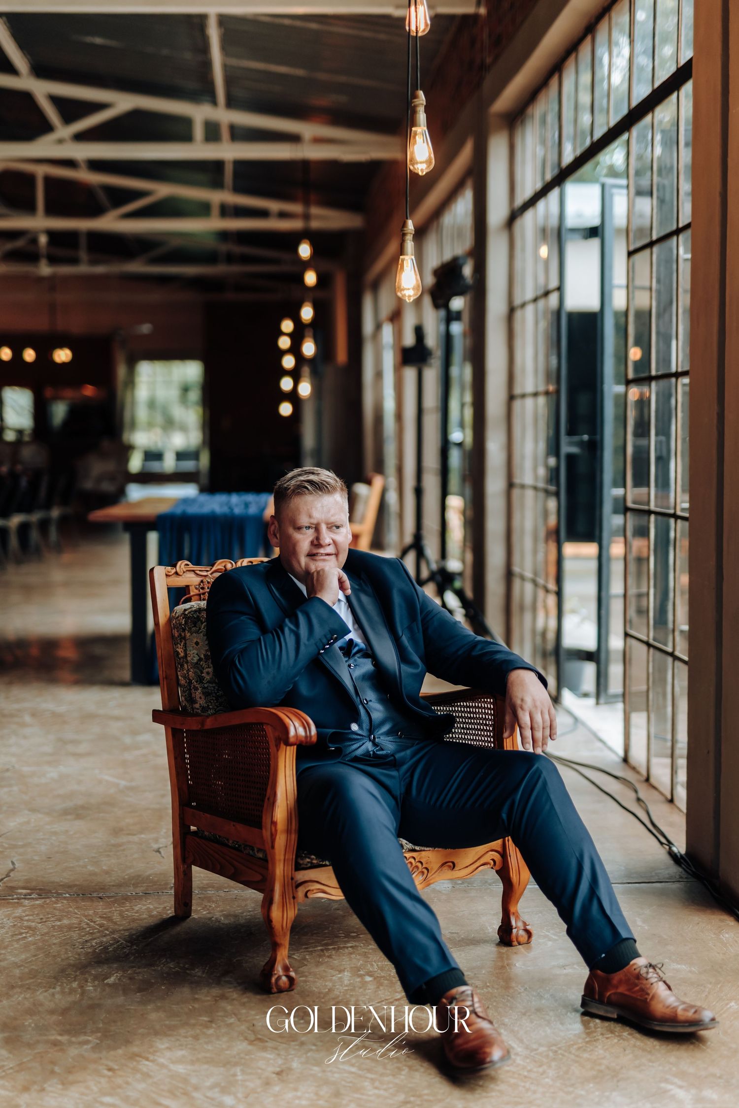 Man in navy suit seated in ornate wooden chair at elegant venue with Edison bulb lighting.