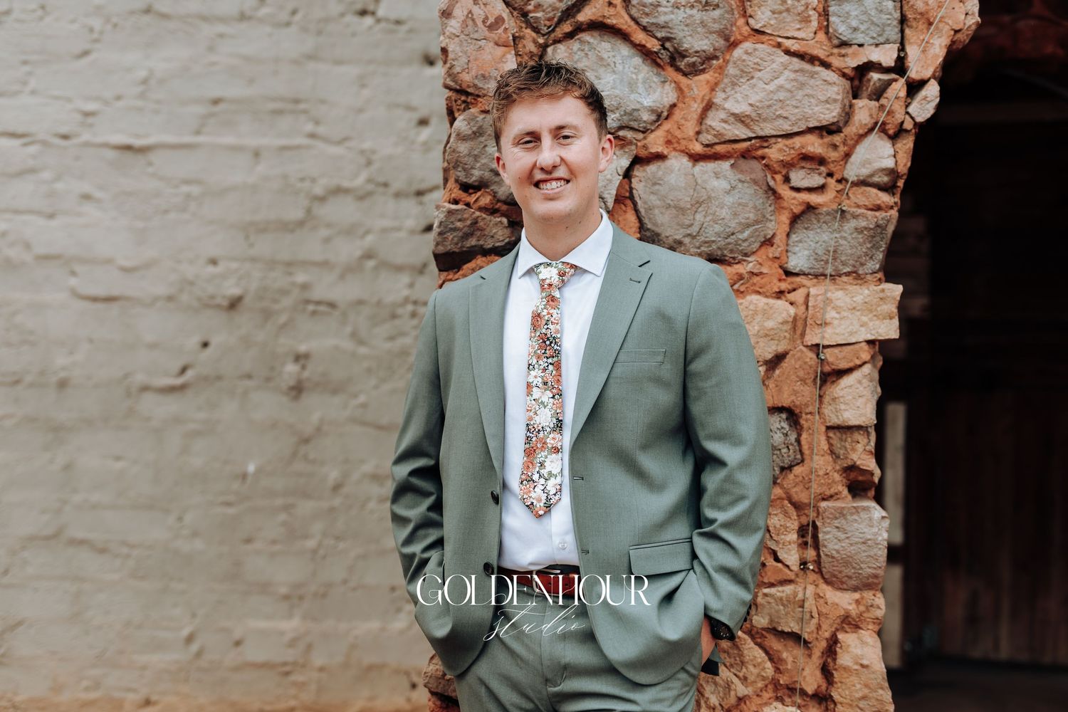 A person in a sage green suit and floral tie poses against a rustic stone wall in three similar photos.