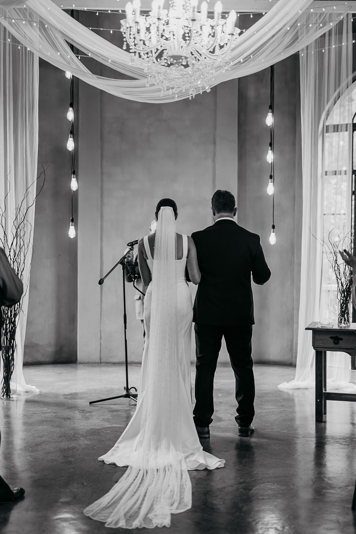 Black and white wedding ceremony silhouette with elegant white draping and hanging lights in an industrial space.