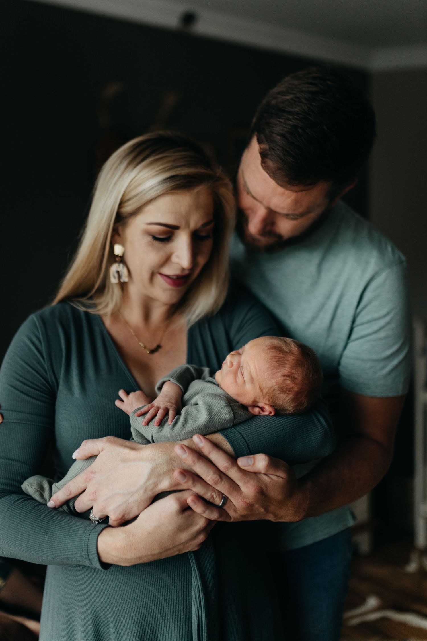 A couple shares an intimate moment with their newborn baby in a darkly lit room.