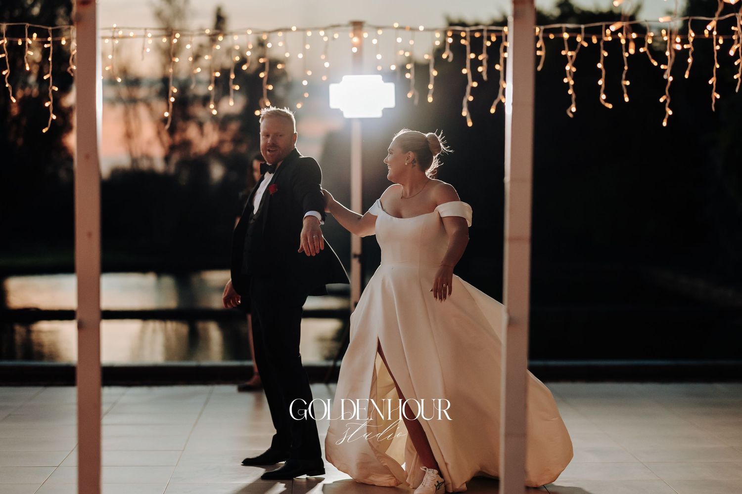 A couple shares their first dance at a wedding reception under twinkling string lights and wooden beams.