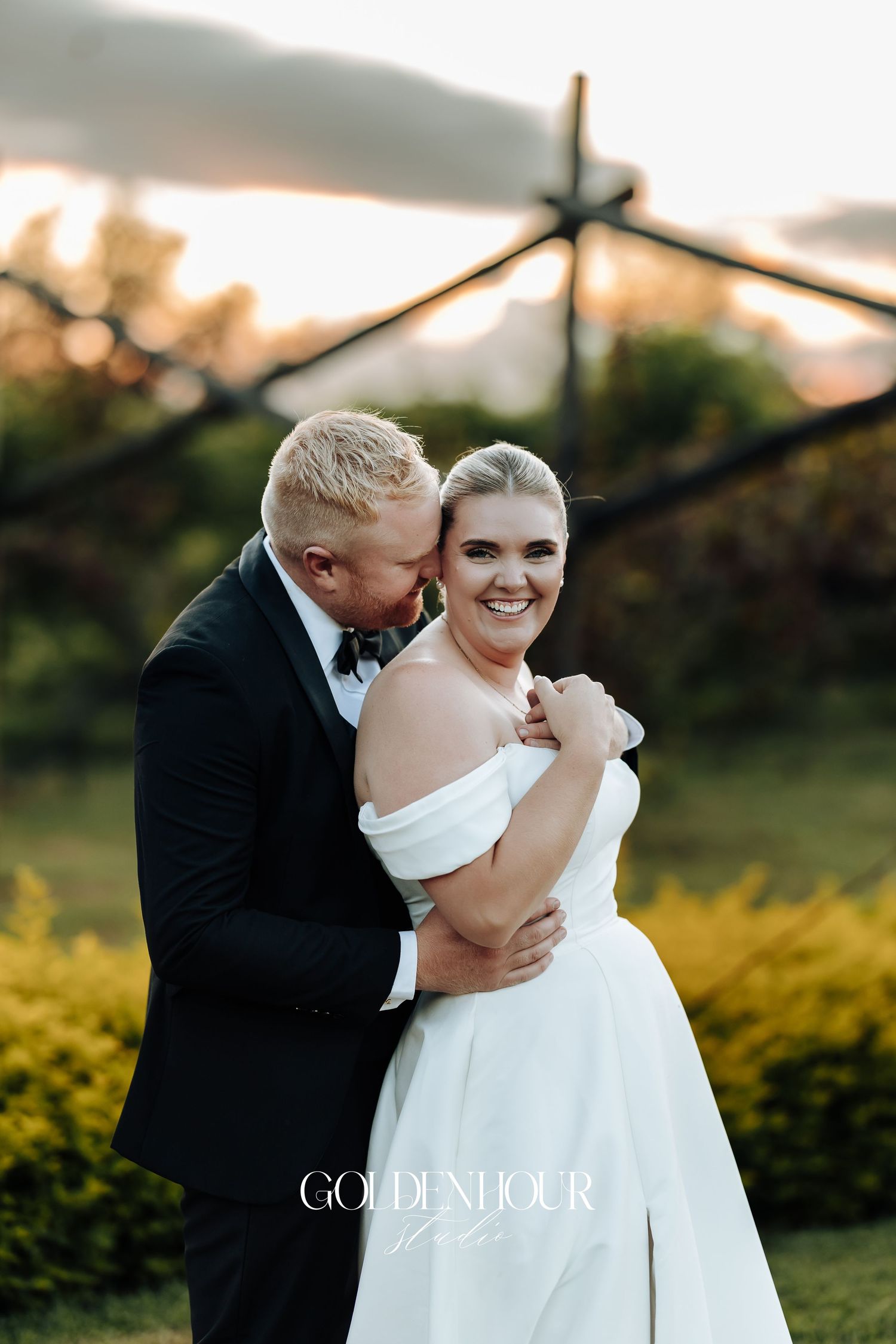 A romantic wedding couple embraces outdoors at sunset with golden light filtering through trees in the background.