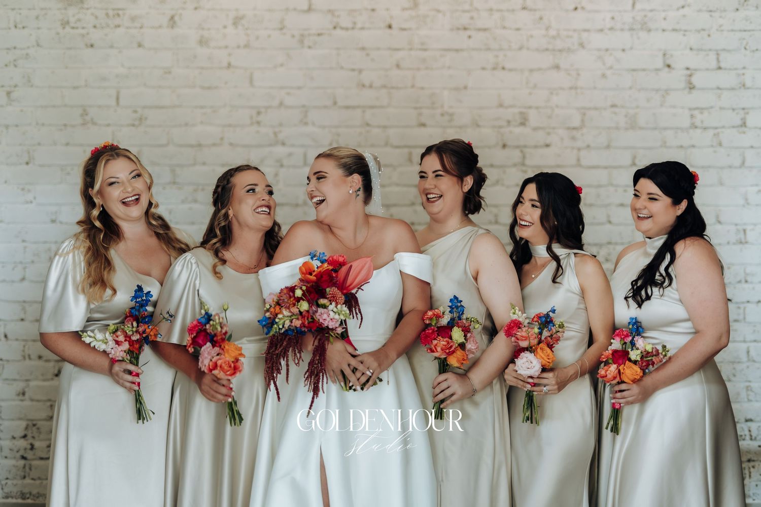 Bridesmaids in champagne colored dresses stand in a row holding vibrant coral bouquets against a white brick wall.
