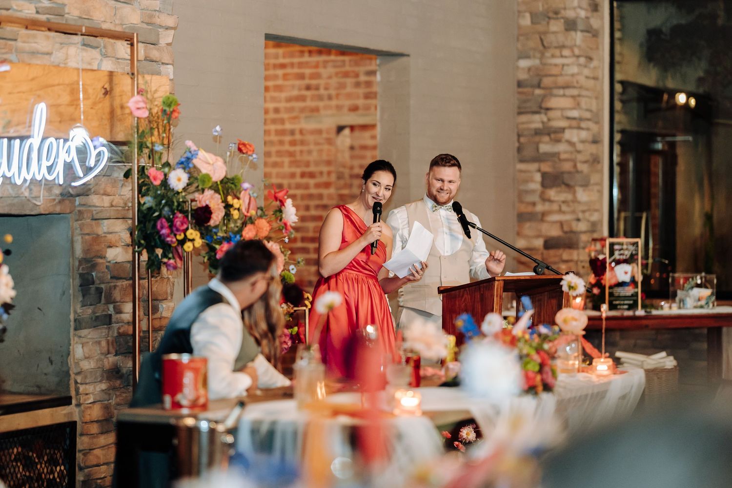 Wedding speech being given at reception with floral decorations and brick wall backdrop.