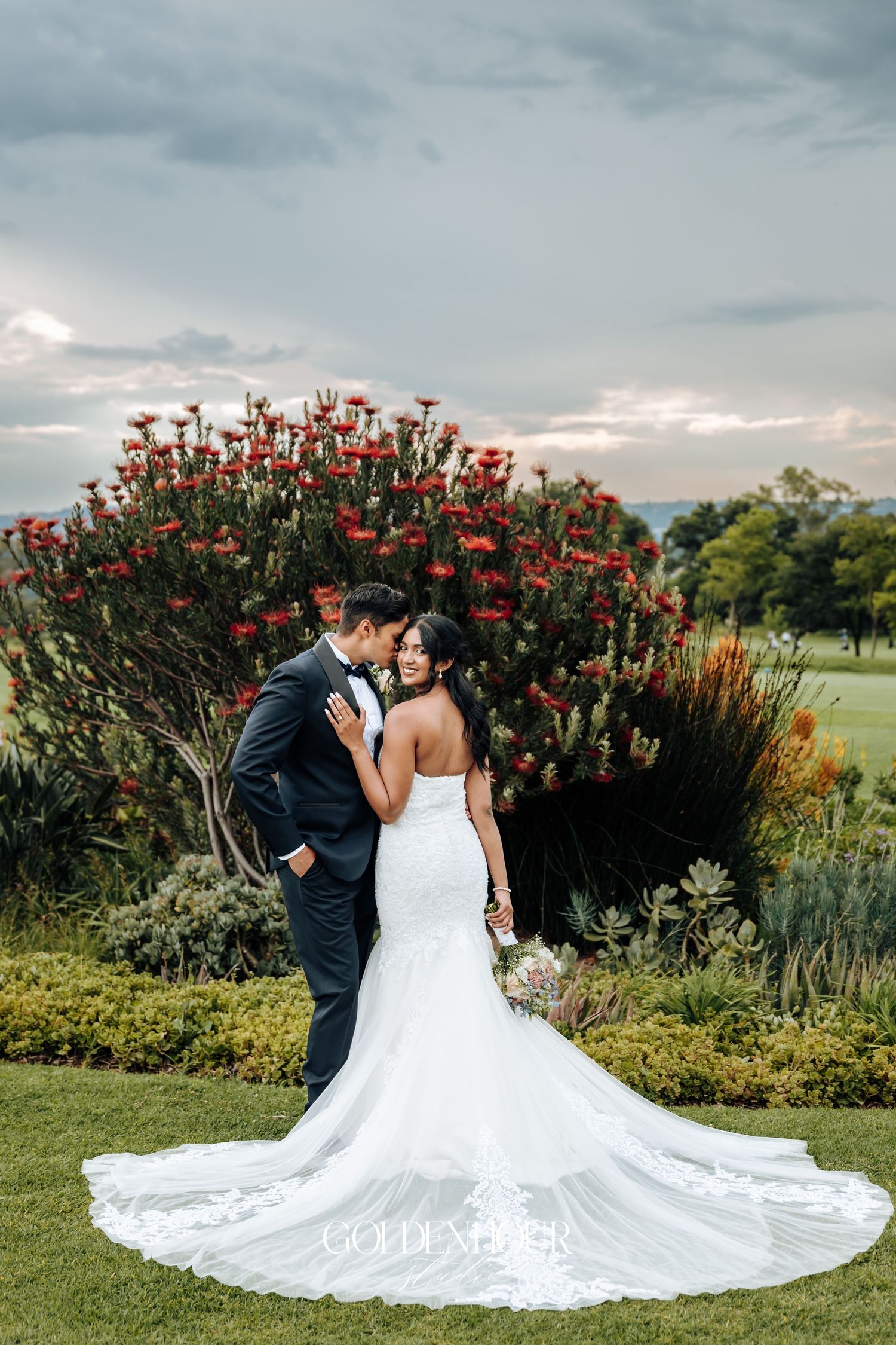 Wedding couple embracing in front of blooming red flowers in garden setting.