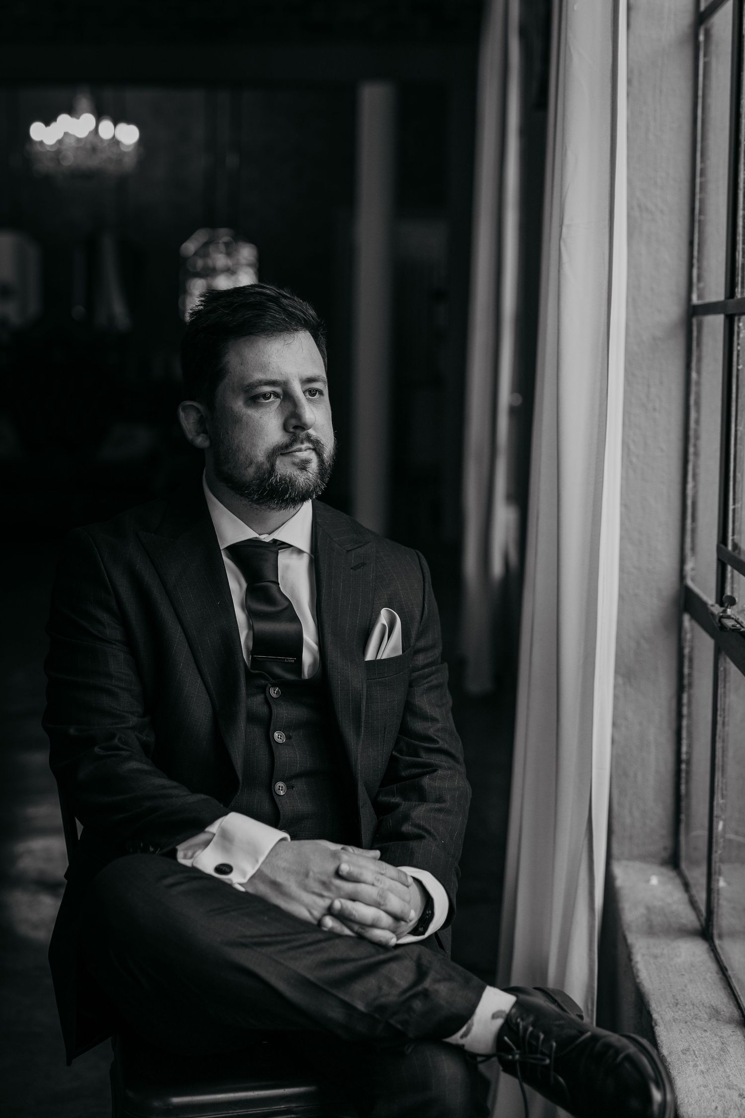 Person in formal attire sitting by window in dramatic black and white portrait.