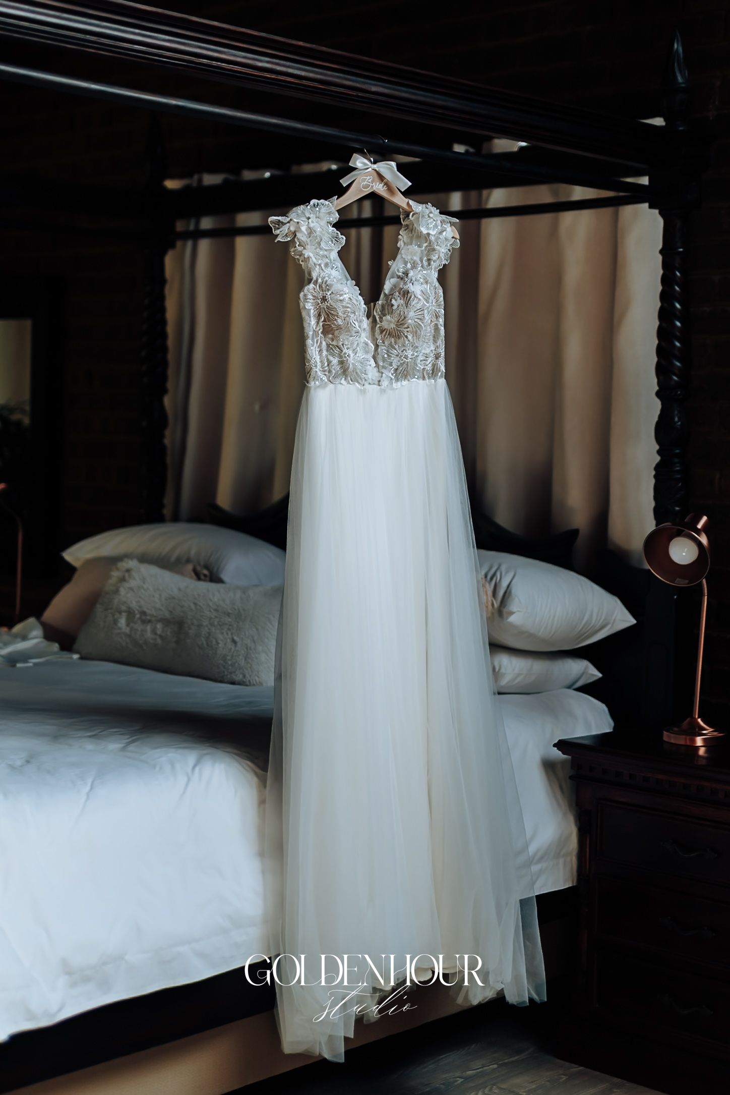 Elegant lace and tulle wedding gown hanging on a wooden hanger above a dark four-poster bed.