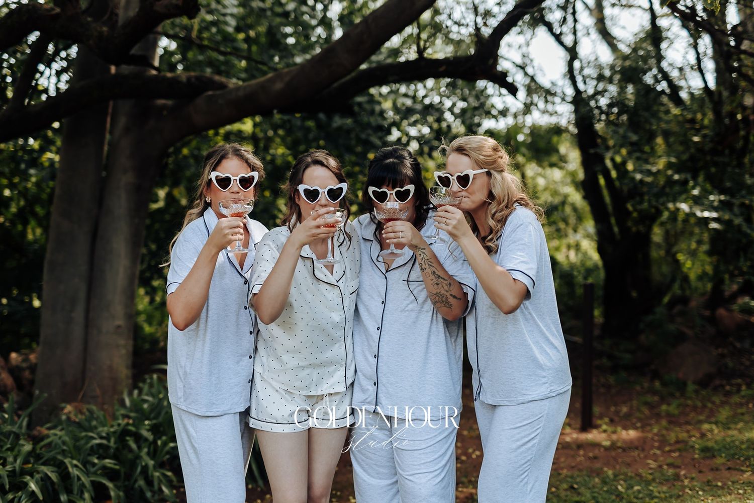 Four bridesmaids in matching pajamas wearing heart-shaped sunglasses posing outdoors before wedding.