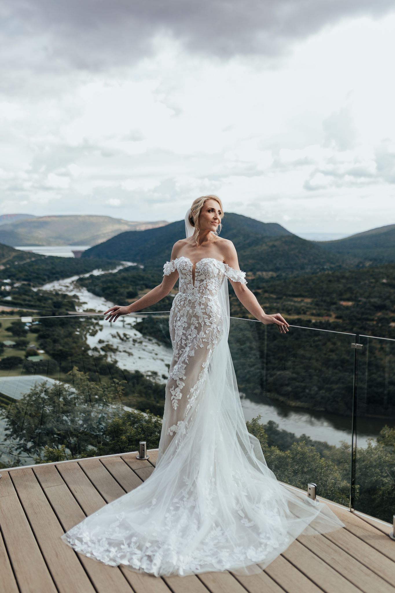 Bride in lace wedding dress poses on deck overlooking dramatic river valley and mountains.