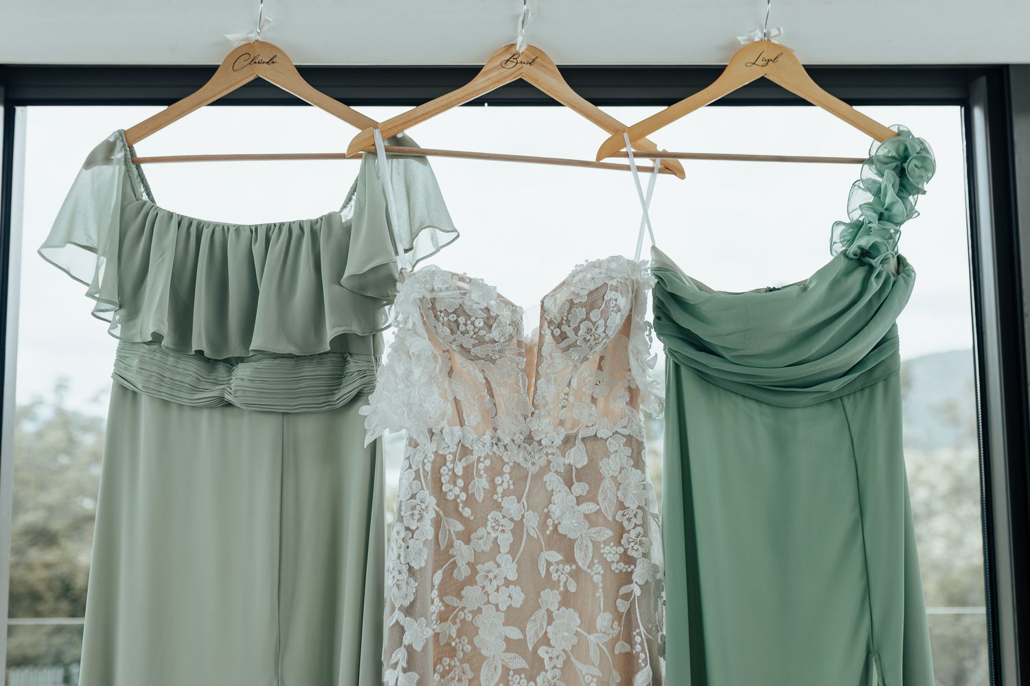 Three bridesmaid dresses in sage green tones hanging beside white lace wedding gown.