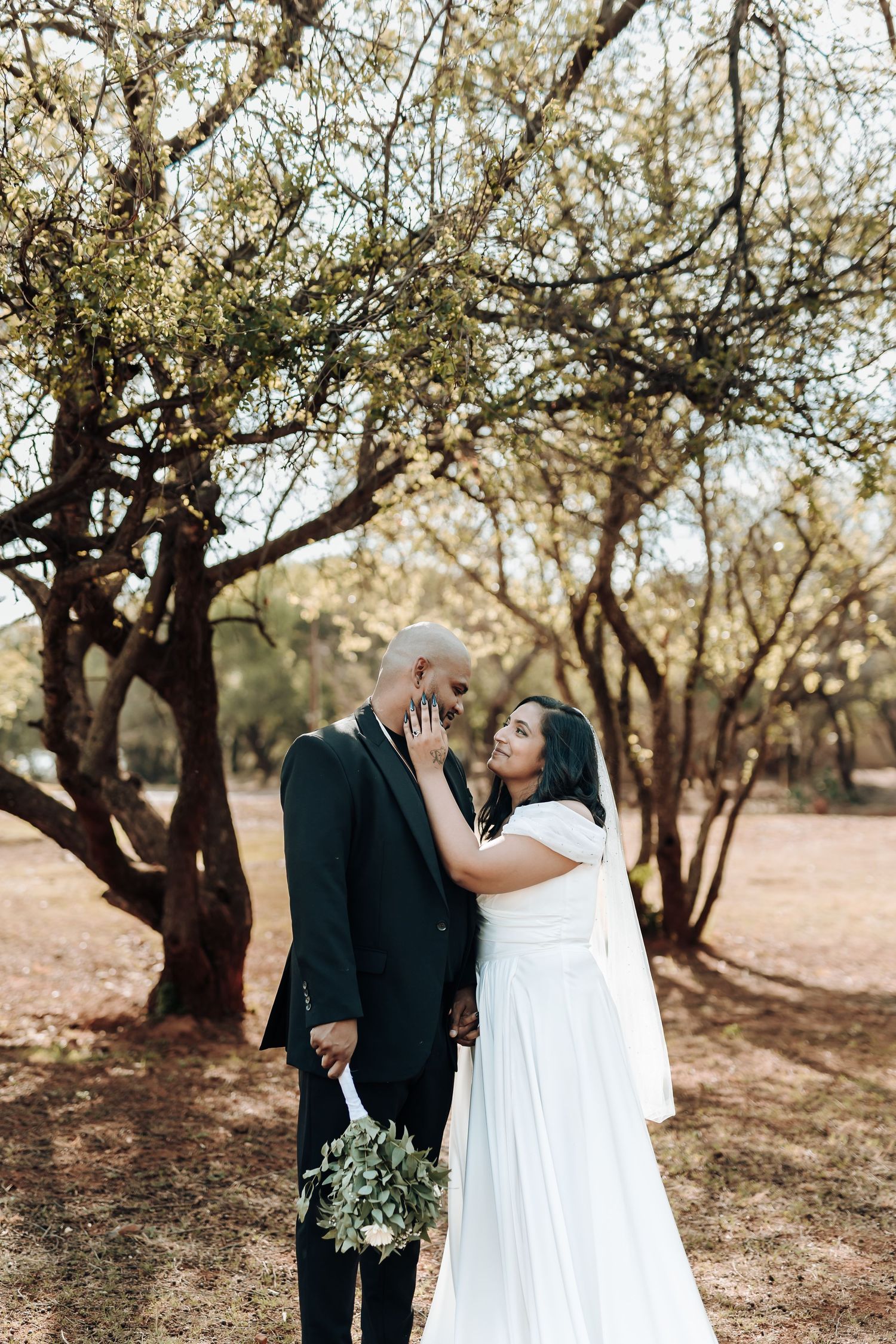 Wedding couple poses intimately among trees with bride holding greenery bouquet.