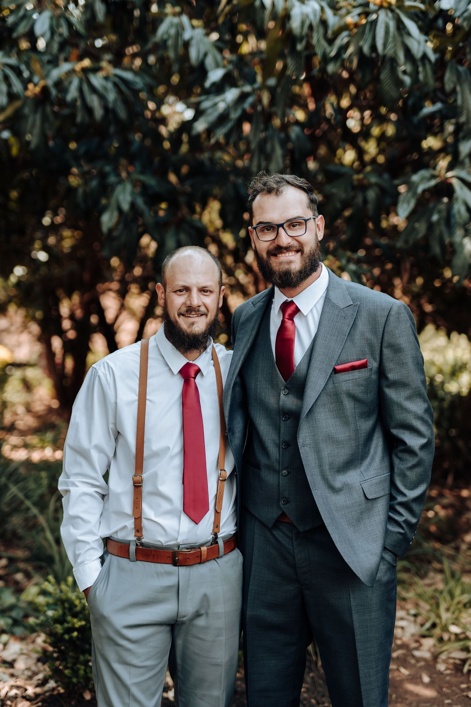 Two men in formal wedding attire posing against autumn foliage background.
