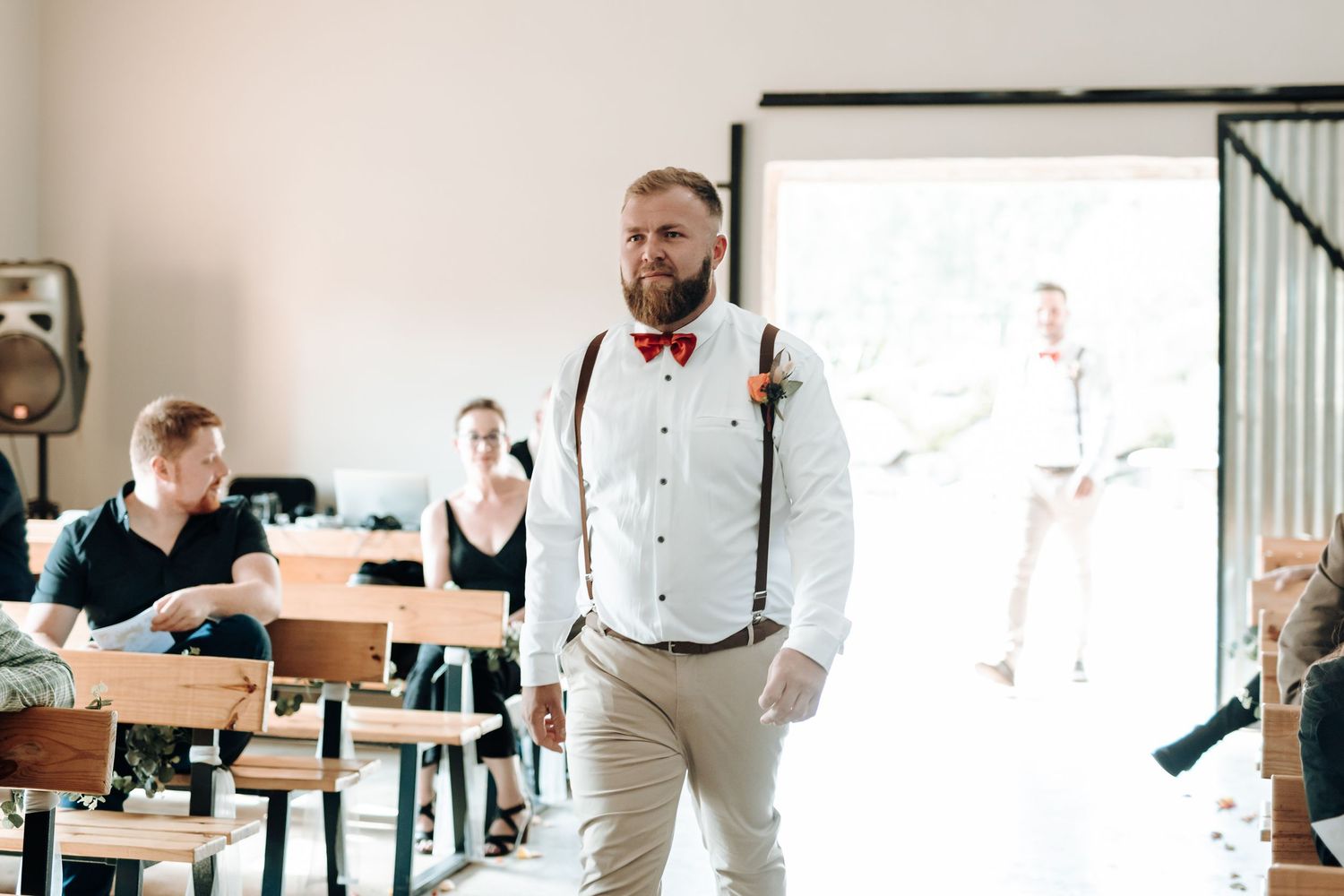 Groom in suspenders and bow tie walks through bright doorway during wedding ceremony.